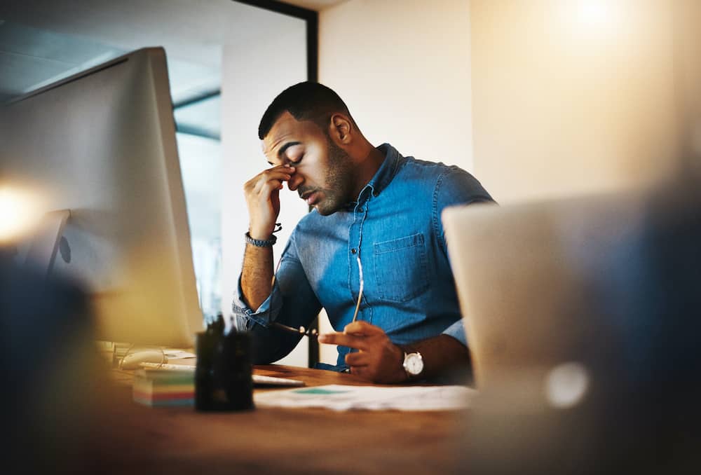 Exhausted man at sitting at desk with hand on head with symptoms of chronic fatigue syndrome