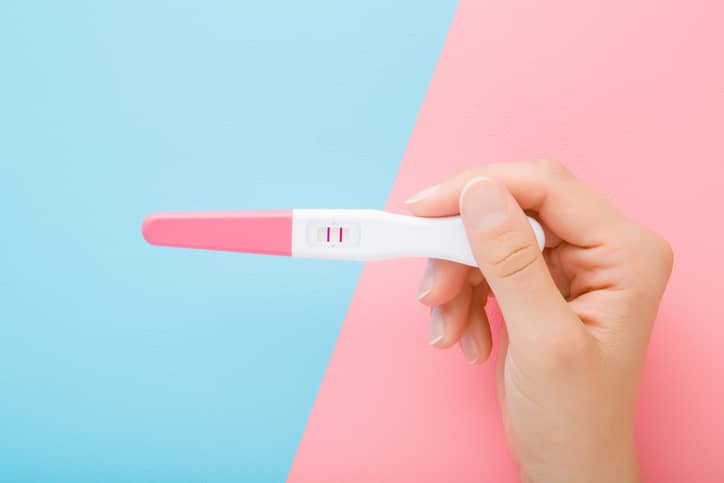 Hand holding pregnancy test with two stripes on light pink blue table background.