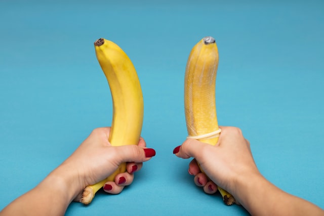 Hands holding 2 bananas 1 with condom 1 without on blue