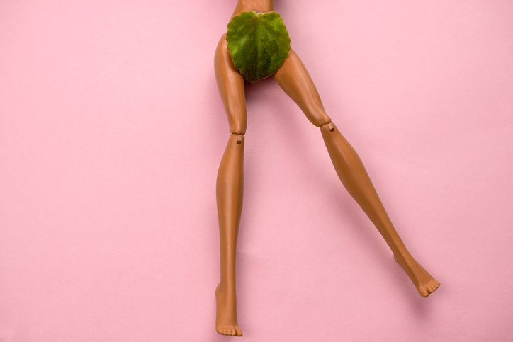 Legs of a plastic doll with a green leaf covering the crotch area on a pastel pink background