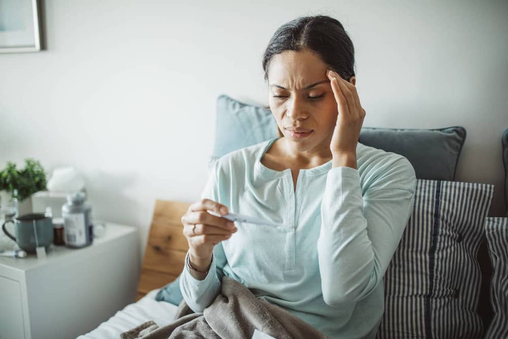 Person sitting up in bed with hand on head measuring temperature using thermometer