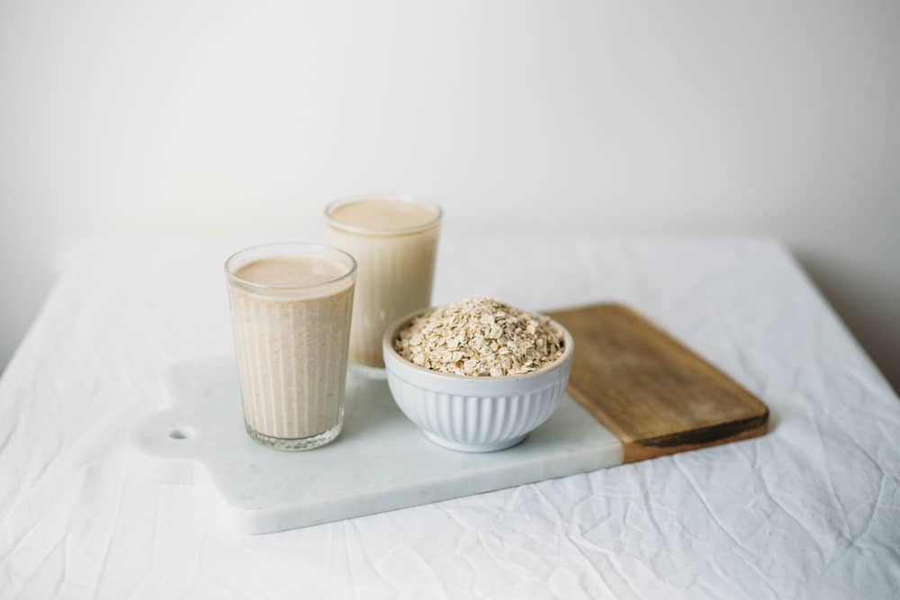 2 glasses of oat milk next to bowel of oats on board