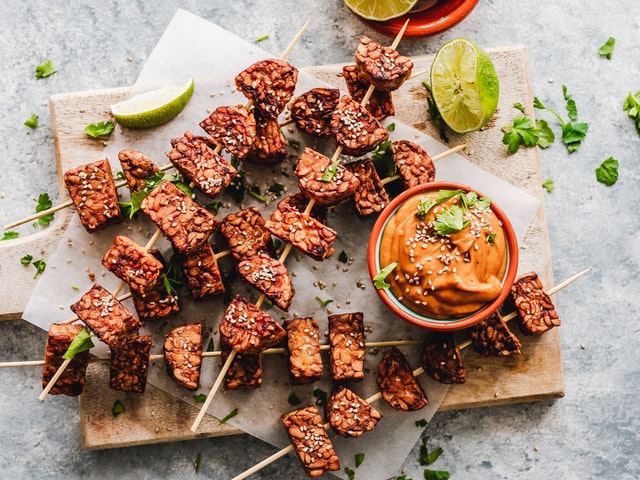 Sesame tempeh skewers on a chopping board with spicy peanut sauce.