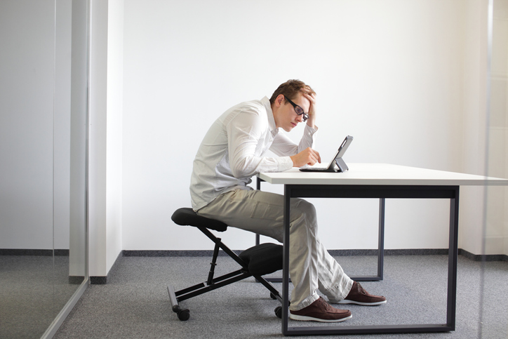 Young man seating on kneeling chair with bad posture at work