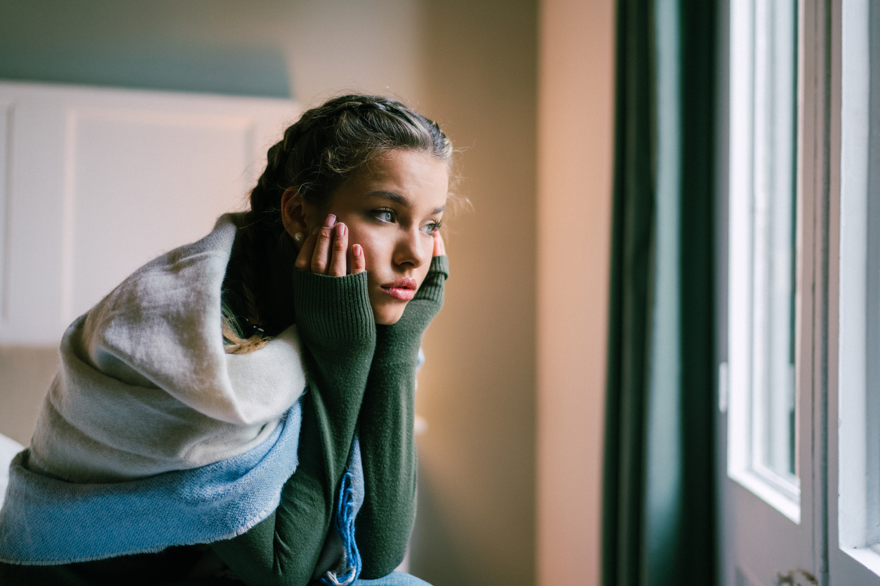 Young woman sitting on a bed looking sad while looking out the window (credit - martin-dm)